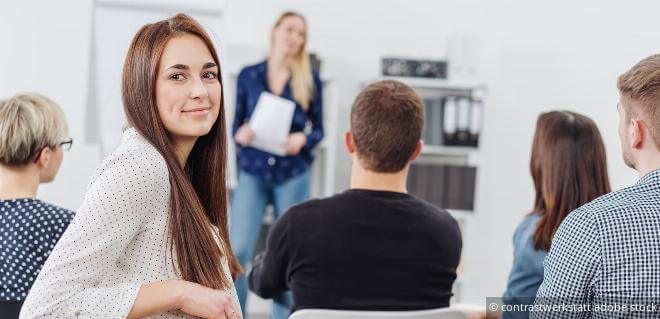 Neue Weiterbildungszeit: Teilnehmerin blickt in die Kamera, während im Hintergrund eine Frau unterrichtet.