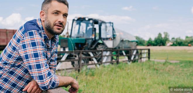 Landwirt lehnt auf einem Zaun mit Mähdrescher im Hintergrund – Symbol für Land- und Forstwirtschaft als Gewerbebetrieb.